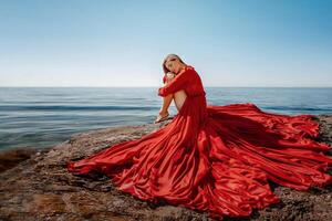 mooi sensueel vrouw in een vliegend rood jurk en lang haar, zittend Aan een rots bovenstaand de mooi zee in een groot baai. foto