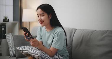 gelukkig jong vrouw zittend Aan sofa gebruik makend van credit kaart met mobiel telefoon voor online boodschappen doen zonder contant geld in leven kamer Bij huis foto