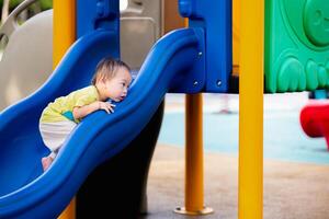 portret van een Aziatisch baby jongen ernstig proberen naar beklimmen blauw schuiven, kleuter spelen met speelgoed- temidden van een buitenshuis speelplaats, kind met heet weer en zweten, kleuter van een jaar en drie maanden oud foto