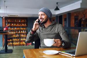 jong knap hipster Mens met baard zittend in cafe pratend mobiel telefoon, Holding kop van koffie en lachend. laptop Aan houten tafel. foto