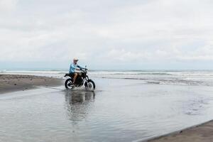 Mens in pet rijden motorfiets Aan strand. moto kruis crossmotor Aan strand zonsondergang Aan Bali. jong hipster mannetje genieten van vrijheid en actief levensstijl, hebben pret Aan een motorrijders tour. foto