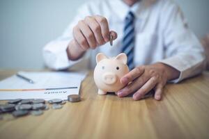 persoon in een bedrijf overhemd besparing geld in een varkentje bank, met munten en financieel rapporten Aan de tafel. besparing geld bedrijf concept foto