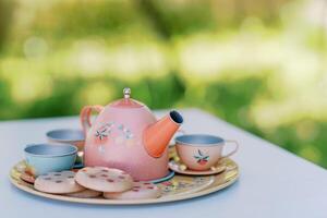 kleurrijk speelgoed- theepot met cups en koekjes staat Aan een dienblad Aan een tafel in de tuin voor een thee partij foto