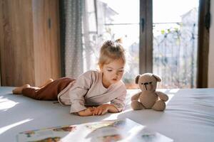 weinig meisje leugens Aan haar kant Aan een bed met een teddy beer en looks Bij een afbeelding boek foto