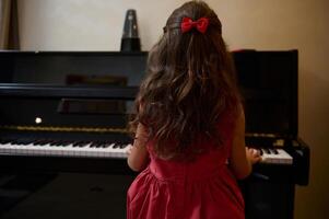 terug visie van een weinig kind meisje in elegant rood jurk, zittend Bij piano sterk, het uitvoeren van klassiek melodie, gevoel ritme van muziek, aanraken zwart en wit ivoor en ebbehout sleutels van de groots piano foto