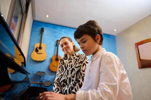 getalenteerd tiener jongen musicus hebben een piano les met zijn docent. vrouw pianist uitleggen de correct positie van handen Aan piano sleutels foto