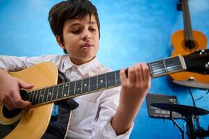 geïnspireerd tiener- jongen verloofd in spelen gitaar, tokkelen de strings van zijn akoestisch musical instrument, tokkelen in musical studio met andere instrumenten, trommels. hobby's en vrije tijd foto