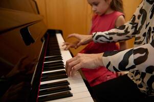 detailopname vrouw pianist Holding handen Aan piano toetsenbord, aanraken zwart en wit sleutels, voorvormen klassiek melodie gedurende terwijl geven een muziek- les naar haar weinig leerling meisje. foto