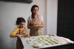 lief weinig meisje in beige chef schort, staand Bij bebloemd keuken tafel, helpen haar moeder naar dingen en beeldhouwen knoedels. foto