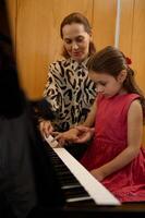 verticaal detailopname portret van een schattig weinig meisje in elegant rood jurk, zittend Bij houten piano, met leraar aan het leren naar Speel piano binnenshuis foto