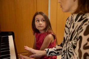 authentiek portret van mooi Kaukasisch elementair leeftijd schoolmeisje in elegant rood jurk, glimlachen op zoek Bij haar muziek- docent, zittend samen Bij piano gedurende individu muziek- les binnenshuis foto