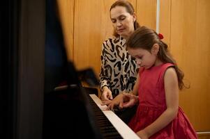 aanbiddelijk weinig meisje in rood jurk, nemen piano les, Met passie spelen de sleutels onder haar leraar de begeleiding, gevoel de ritme van muziek. musical onderwijs en talent ontwikkeling in vooruitgang foto