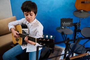 spaans tiener- jongen in wit overhemd en blauw jeans, zittend in zijn blauw studio kamer, spelen akoestisch gitaar, gefocust Aan zijn muziek, vulling de lucht met de geluiden van zijn instrument foto