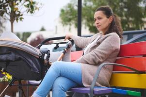 jong vrouw zittend Aan bank geschilderd in regenboog kleuren, wandelen met haar weinig pasgeboren kind slapen in een baby kinderwagen foto