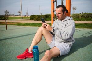 jong atletisch Mens zittend Aan de sport- grond en controle Gezondheid sport mobiel app Aan smartphone na buitenshuis training foto