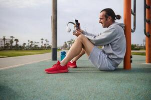 atletisch Mens controle mobiel app Aan zijn smartphone terwijl resting na training de stedelijk sport- grond. kant visie foto