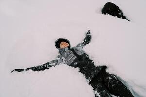 jong glimlachen vrouw in een ski pak leugens in de sneeuw met haar armen uitgestrekt in de buurt een zwart hond foto