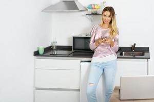 blonde vrouw die haar smartphone gebruikt die thuis in de keuken staat