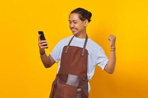 portret van een aziatische jongeman met een schort die er opgewonden uitziet en succes viert met opgeheven handen en een smartphone vasthoudt foto