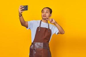 Portret van een Aziatische man verrast en toont vredesteken terwijl hij selfie neemt op mobiel geïsoleerd op gele achtergrond foto