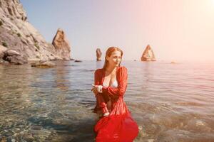 vrouw reizen zee. gelukkig toerist in rood jurk genieten nemen afbeelding buitenshuis voor herinneringen. vrouw reiziger poseren in zee strand, omringd door vulkanisch bergen, sharing reizen avontuur reis foto