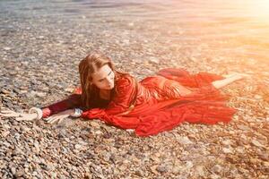 vrouw reizen zee. gelukkig toerist in rood jurk genieten nemen afbeelding buitenshuis voor herinneringen. vrouw reiziger poseren in zee strand, omringd door vulkanisch bergen, sharing reizen avontuur reis foto