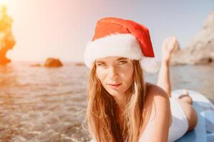 vrouw zee sup. dichtbij omhoog portret van gelukkig jong Kaukasisch vrouw met lang haar- in de kerstman hoed op zoek Bij camera en lachend. schattig vrouw portret in een wit bikini poseren Aan sup bord in de zee foto