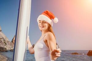 vrouw zee sup. dichtbij omhoog portret van gelukkig jong Kaukasisch vrouw met lang haar- in de kerstman hoed op zoek Bij camera en lachend. schattig vrouw portret in een wit bikini poseren Aan sup bord in de zee foto