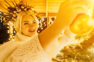 een blond vrouw in wit jurk en een kroon van goud ornamenten versieren Kerstmis boom met goud ornamenten en lichten. de boom is versierd met goud ballen en is lit omhoog met lichten. foto