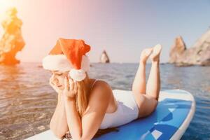 vrouw zee sup. dichtbij omhoog portret van gelukkig jong Kaukasisch vrouw met lang haar- in de kerstman hoed op zoek Bij camera en lachend. schattig vrouw portret in een wit bikini poseren Aan sup bord in de zee foto