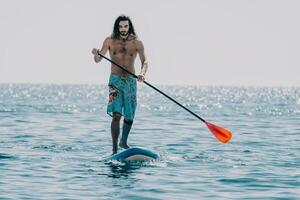 Mens sup zee. sterk atletisch Mens leert naar peddelen sup staand Aan bord in Open zee oceaan Aan zonnig dag. zomer vakantie vakantie en reizen concept. antenne visie. langzaam beweging foto