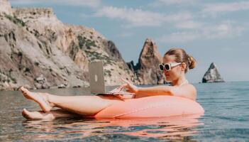 vrouw freelancer werken Aan laptop zwemmen in zee Aan roze opblaasbaar ring. gelukkig toerist in zonnebril drijvend Aan opblaasbaar donut en werken Aan laptop computer in kalmte oceaan. afgelegen werken overal foto