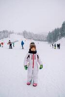 weinig glimlachen meisje staat Aan de ski helling tegen de backdrop van skiërs foto