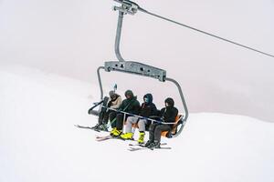 skiërs met ski polen in hun handen rijden een vierzitter stoeltjeslift omhoog een besneeuwd berg helling foto