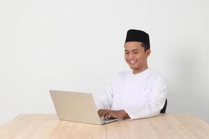 portret van opgewonden Aziatisch moslim Mens in koko overhemd met kalotje werken Aan zijn laptop gedurende vastend Aan Ramadan maand. geïsoleerd beeld Aan wit achtergrond foto