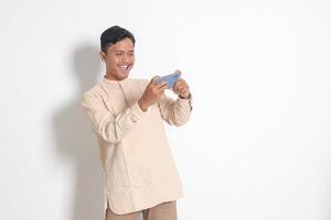 portret van jong opgewonden Aziatisch moslim Mens in koko overhemd Holding mobiel telefoon en spelen spellen Aan zijn smartphone. geïsoleerd beeld Aan wit achtergrond foto