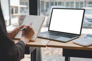 persoon is ijverig schrijven in een notitieboekje naast een strak laptop, met een rekenmachine Bij hand, wijzend op een focus Aan bedrijf planning, budgetteren, of financieel analyse in een helder, modern werkruimte. foto