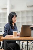 glimlachen Aziatisch vrouw gefocust Aan haar laptop werk in een helder cafe milieu met koffie en schrijfbehoeften Aan de tafel. foto