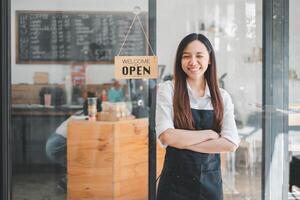 gastvrij cafe eigenaar met armen gekruist, glimlachen in voorkant van haar bedrijf met een Open teken, afbeelden ondernemerschap en klant onderhoud. foto