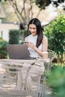 jong vrouw glimlachen net zo ze toepassingen haar smartphone, met een laptop Open in voorkant van haar Aan een tafel in een buitenshuis tuin instelling. foto