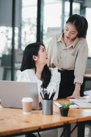 twee professioneel Dames bezighouden in een samenwerkend discussie Bij een goed georganiseerd kantoor bureau, met een zittend en de andere staan, sharing een moment van vriendelijk interactie. foto