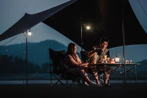 net zo nacht valt, een paar smaken hun koffie en geniet intiem gesprek Bij hun goed verlicht oever van het meer camping. foto