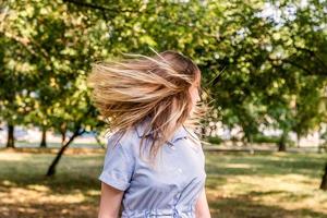 Jonge blanke blonde vrouw in een blauwe gestripte zomerjurk schudt haar haar in het park buiten op een zonnige dag foto