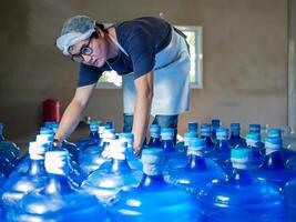 Aziatisch Mens arbeider of kwaliteit inspecteur in werkkleding werken in controle drinken water blauw gallon in drinken water fabriek voordat zending.drinken water zakelijk, klein bedrijf, winkel, magazijn foto