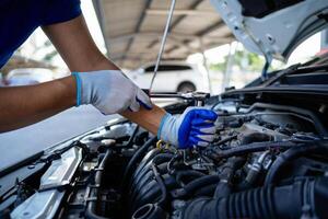 een Mens is werken Aan een auto motor. hij is vervelend blauw handschoenen en een blauw shirt. de auto is geparkeerd onder een dak foto