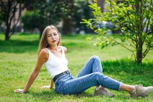 mooi jong vrouw vervelend tank top en jeans zittend Aan gras gazon foto