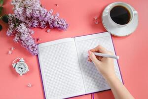 voorjaar plat leggen. kladblok, lila bloemen en kop van koffie Aan koraal achtergrond. foto