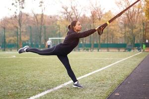 meisje atleet functioneel opleiding Aan sport veld. gemengd ras jong volwassen vrouw Doen training met suspensie systeem. gezond levensstijl. uitrekken buitenshuis speelplaats. foto