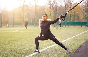 meisje atleet functioneel opleiding Aan sport veld. gemengd ras jong volwassen vrouw Doen training met suspensie systeem. gezond levensstijl. uitrekken buitenshuis speelplaats. foto