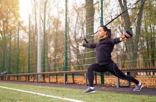 meisje atleet functioneel opleiding Aan sport veld. gemengd ras jong volwassen vrouw Doen training met suspensie systeem. gezond levensstijl. uitrekken buitenshuis speelplaats. foto